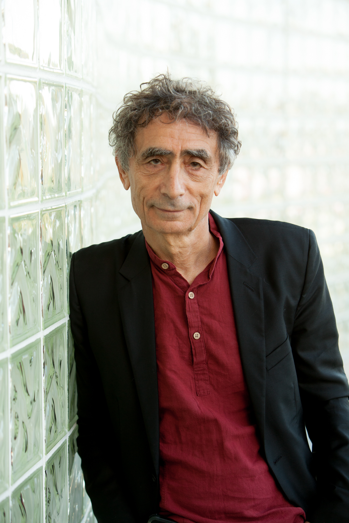 Op de afbeelding is Gabor Maté te zien, de auteur van 'Hongerige geesten'. Het is een witte man van 78 jaar. Hij heeft peper-en-zoutkleurig haar. Hij draagt een rode blouse met daaroverheen een zwarte colbert. 